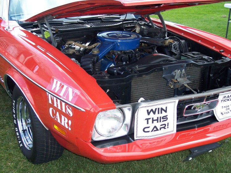 Red Ford Mustang, hood up showing engine. "WIN THIS CAR!" signs on fender and grille
