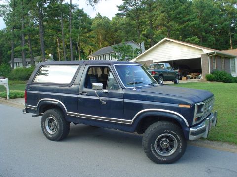 1984 Ford Bronco - A dark blue SUV is parked on the street in front of a house with a garage containing another vehicle.