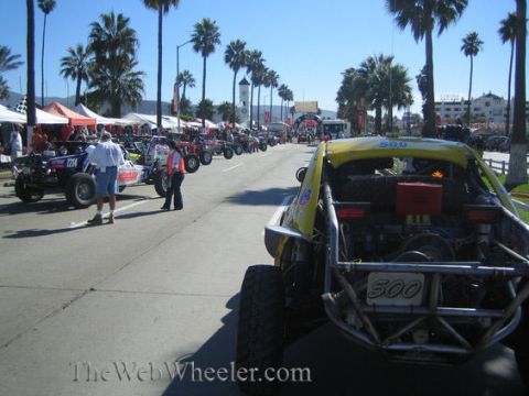 1990 Ford Bronco - yellow tube-frame off-road race car numbered 500 sits at a palm-lined coastal staging area among other race vehicles and crew.