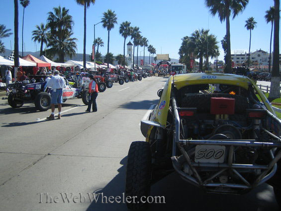 Yellow off-road race buggy #500 at staging area lined with palm trees and tents