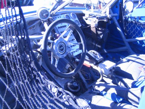1990 Ford Bronco - cockpit of an off-road race car shows a polished steering wheel, gauge cluster, roll cage tubing and a mesh window net.