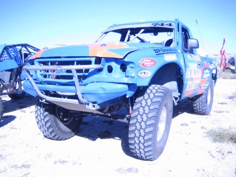 1990 Ford Bronco - blue and orange off-road race truck numbered 744 with a stripped front bumper and large mud tires sits on desert ground.