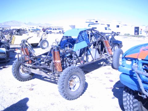 1990 Ford Bronco - open tube-frame off-road buggy with orange coilover shocks and big tires sits in a dusty desert pit area near RVs.