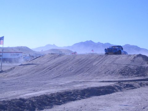 1990 Ford Bronco - blue race truck crests a graded dirt berm with an American flag and desert mountains in the background.