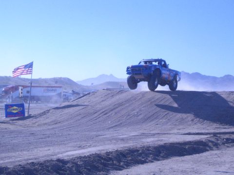 1990 Ford Bronco - blue off-road race truck launches airborne over a dirt jump as dust trails behind, with an American flag and mountains beyond.