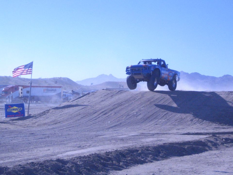 Blue off-road race truck airborne over a dirt jump with American flag in background