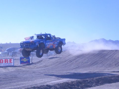 1990 Ford Bronco - blue Horizon Realty off-road race truck flies over a dirt jump with all four wheels off the ground in a desert race.