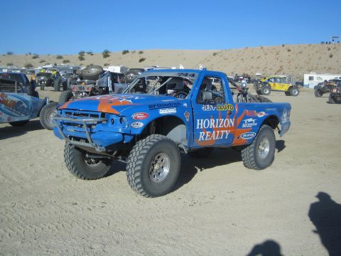 1990 Ford Bronco - blue and orange Horizon Realty off-road race truck numbered 744 sits on desert dirt amid other race vehicles and RVs.