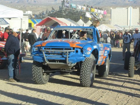 1990 Ford Bronco - blue and orange off-road race truck numbered 744 moves through a crowded pit area with spectators, tents and grandstands behind.