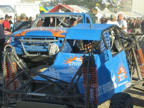 1990 Ford Bronco - blue Horizon Realty tube-frame buggy numbered 218 sits beside a blue and orange race truck among a large pit-area crowd.