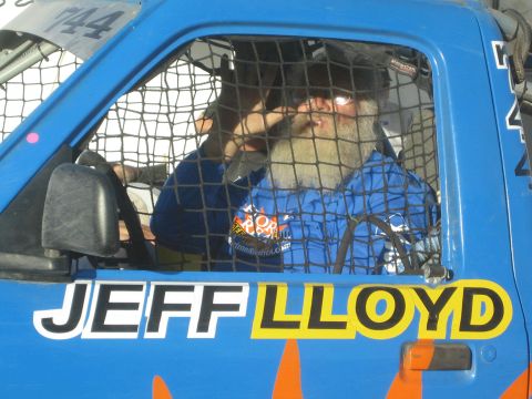 1990 Ford Bronco - bearded driver in a blue racing suit sits behind a mesh window net in a blue race truck lettered JEFF LLOYD on the door.