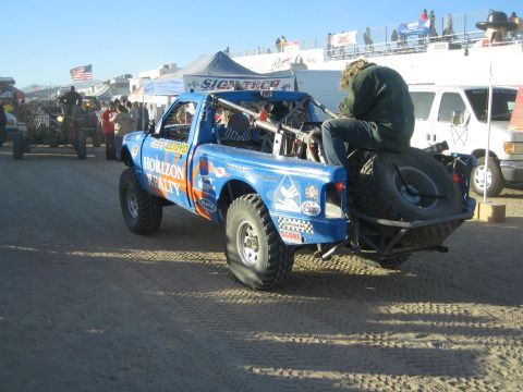 1990 Ford Bronco - blue Horizon Realty off-road race truck with a rear spare tire and a person riding on back drives through a busy desert event area.
