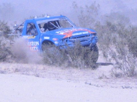 1990 Ford Bronco - blue Horizon Realty off-road race truck powers through desert scrub, throwing up a cloud of dust during a race.