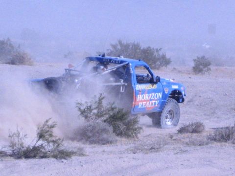 1990 Ford Bronco - blue Horizon Realty off-road race truck slides through open desert terrain trailing a plume of dust.