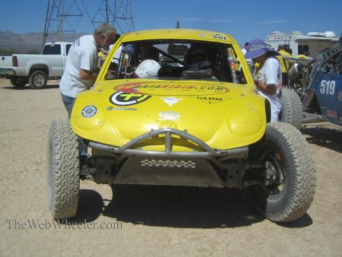 1990 Ford Bronco - front of a yellow Volkswagen Beetle-bodied off-road race car numbered 501 with a tubular bumper, attended by crew in the desert pits.