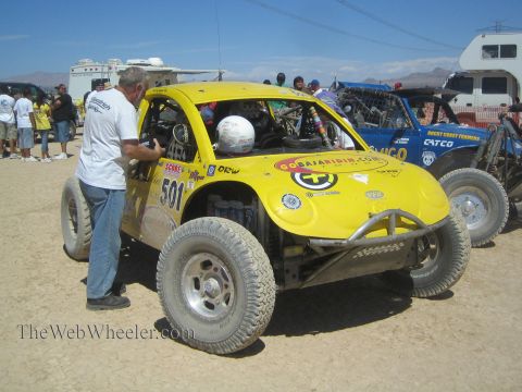 1990 Ford Bronco - yellow Volkswagen Beetle-bodied off-road race car numbered 501 with a helmeted driver inside while a crewman leans in, in a desert pit.