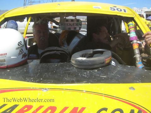 1990 Ford Bronco - Two men sit in the cockpit of a yellow off-road race car numbered 501, one holding a helmet, behind a dust-coated windshield frame.