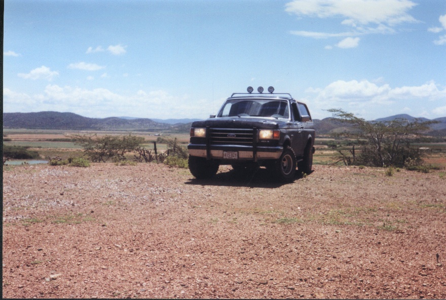 Dark Ford SUV with brush guard and roof light bar parked on gravel in an arid landscape.