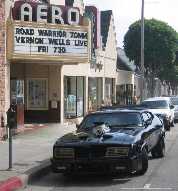 Black 1972 Ford Ranchero with a prominent supercharger, parked on a street in front of a cinema