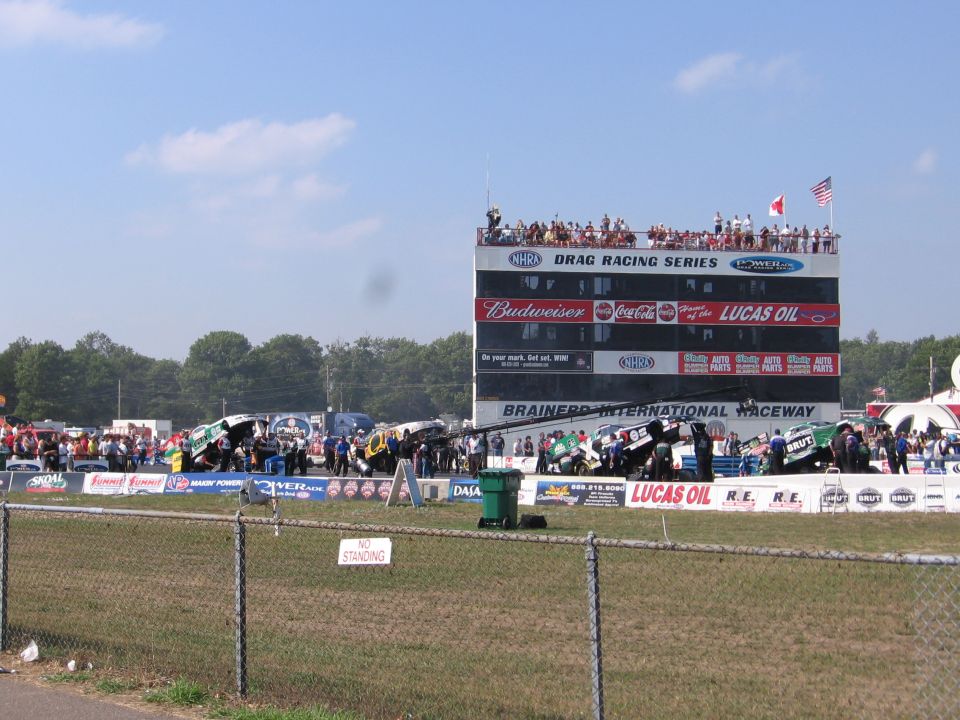 Drag racing track with multiple dragsters lined up, grandstands full of spectators, and a large scoreboard tower