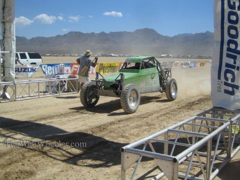 1990 Ford Bronco - green single-seat off-road buggy kicks up dust on a desert course, with a flagger and BFGoodrich and Suzuki banners nearby.