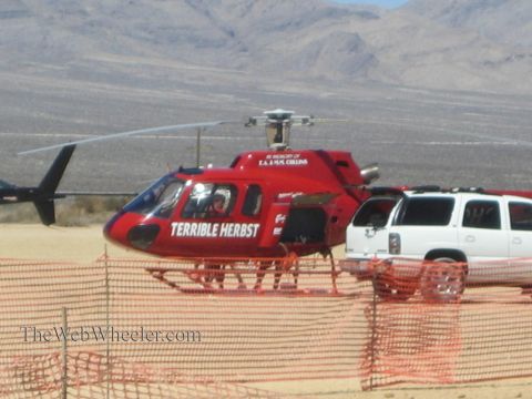 1990 Ford Bronco - red helicopter marked Terrible Herbst sits on desert ground next to a white SUV behind orange safety fencing.