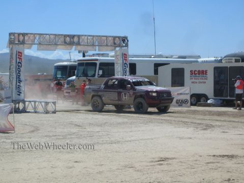 1990 Ford Bronco - Honda off-road race truck passes the SCORE International media center and motorhomes in a dusty desert pit area.