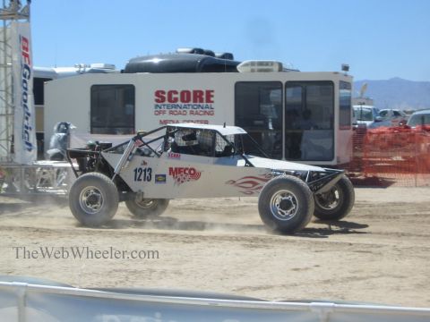 1990 Ford Bronco - white off-road buggy numbered 1213 with flame graphics races past the SCORE International media center trailer.