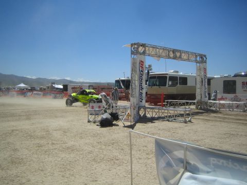 1990 Ford Bronco - green off-road buggy idles near a BFGoodrich truss start gate among motorhomes in a dusty desert paddock.