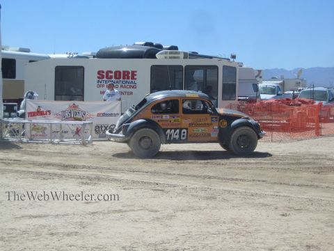 1990 Ford Bronco - orange Baja Bug race car numbered 1148 drives through the desert pit area past the SCORE media center.
