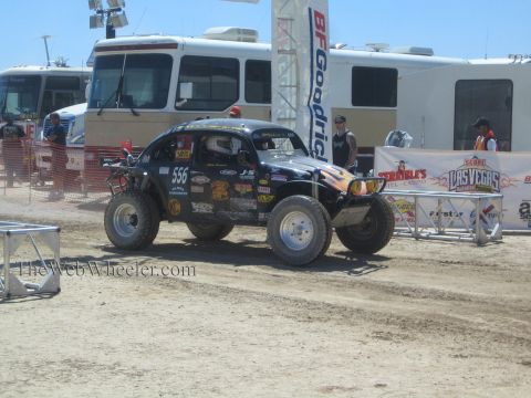 1990 Ford Bronco - black Baja Bug race car numbered 556 drives through the dusty paddock past BFGoodrich and Las Vegas banners.