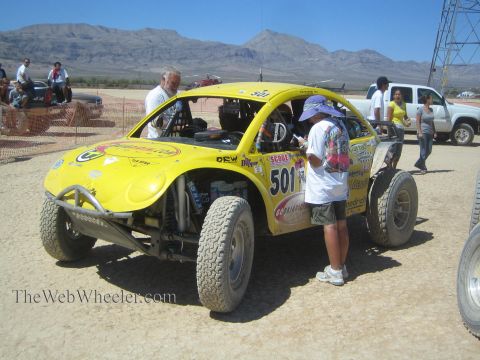 1990 Ford Bronco - yellow Baja Bug race car numbered 501 sits in a desert pit while crew and helpers gather around it.