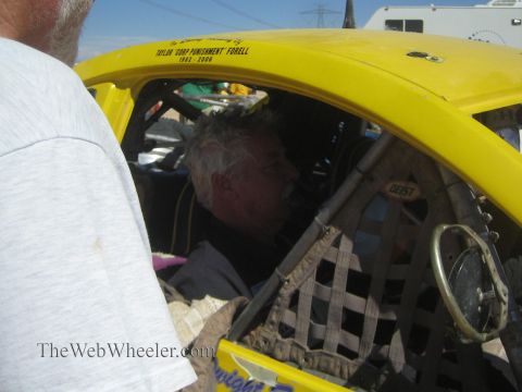 1990 Ford Bronco - gray-haired man sits in the caged cockpit of a yellow Baja Bug, seen past a bystander's shoulder.