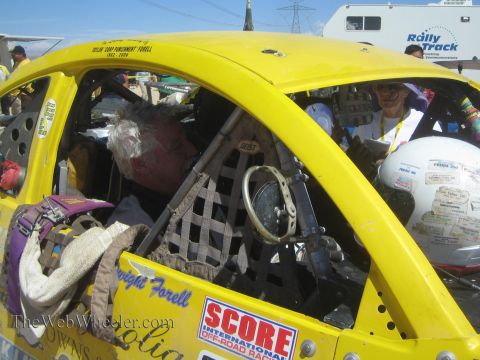 1990 Ford Bronco - gray-haired driver sits inside a yellow Baja Bug's caged cockpit bearing a SCORE International decal in the desert pit.