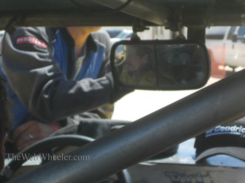 1990 Ford Bronco - crew member in a Pyrotech jacket leans into an off-road race car's cage, his face reflected in the side mirror.