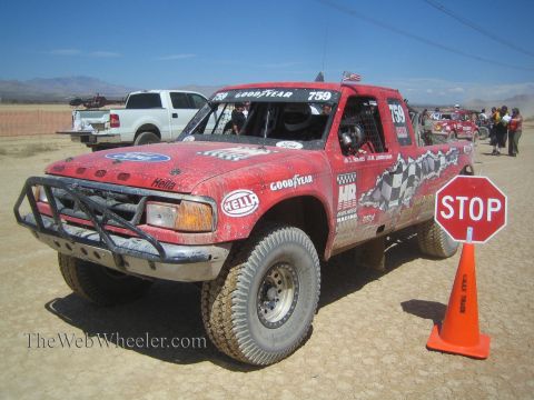 1990 Ford Bronco - red off-road race truck numbered 759 with checkered graphics waits at a desert checkpoint beside a STOP sign and cone.