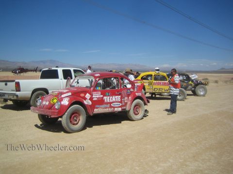 1990 Ford Bronco - red Baja Bug marked Tecate and a yellow off-road buggy stage on a desert road with a pickup and spectators.