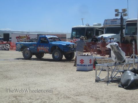 1990 Ford Bronco - blue Horizon Realty off-road race truck sits in a dusty staging area beside Suzuki banners, an RV, and a referee in a striped shirt.