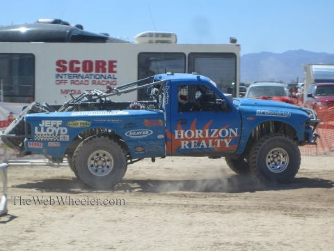 1990 Ford Bronco - blue Jeff Lloyd / Horizon Realty off-road race truck kicks up dust in front of a SCORE International Off Road Racing banner.