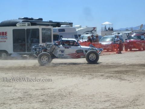 1990 Ford Bronco - white and blue tube-frame off-road buggy numbered 207 races across a dusty course past trailers and orange fencing.
