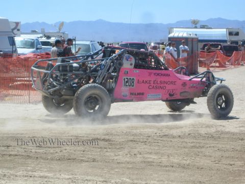 1990 Ford Bronco - pink Lake Elsinore Casino tube-frame off-road buggy numbered 1208 races across the desert past RVs and orange fencing.