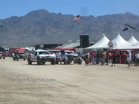 1990 Ford Bronco - Off-road race trucks line up on a dusty desert course beside white tents, spectators and an American flag below distant mountains.
