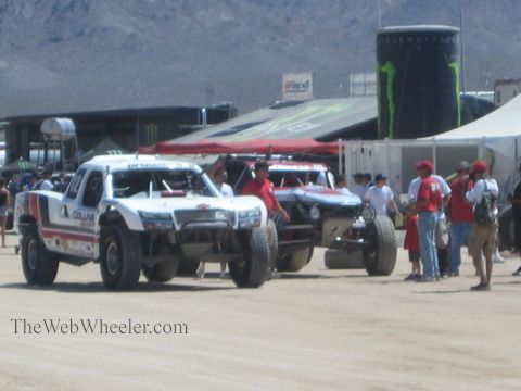 1990 Ford Bronco - White off-road race trucks roll through a dusty pit area past Monster Energy displays, red canopies and a crowd of spectators.
