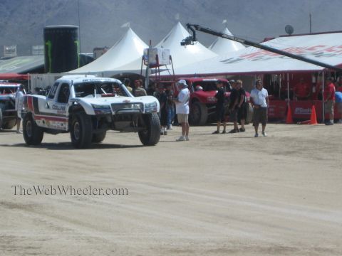 1990 Ford Bronco - white Collins off-road race truck drives along a dusty course past white tents, spectators and a TV camera crane.