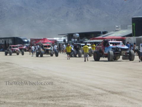 1990 Ford Bronco - Off-road race trucks and buggies stage in a dusty pit area as spectators walk among Monster Energy and Yokohama transporters below mountain…