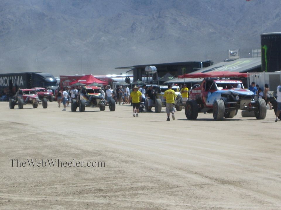 Off-road race trucks and buggies staged in a dusty pit with spectators and Monster Energy rigs