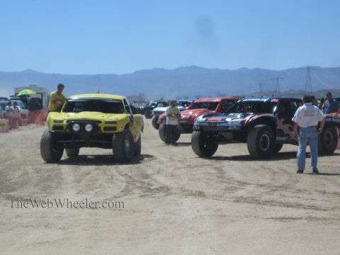 1990 Ford Bronco - yellow off-road race truck and red and dark trucks line up on a dusty desert course as crew members stand nearby below the mountains.