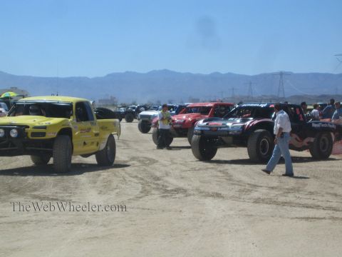 1990 Ford Bronco - row of off-road race trucks, including a yellow one, stages on a dusty course with crew walking and power-line towers in the distance.