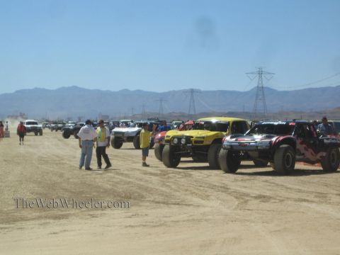 1990 Ford Bronco - long line of off-road race trucks stages on a dusty desert course as crew and spectators gather beneath power-line towers and mountains.