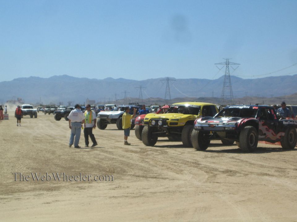 Long line of off-road race trucks staged on dirt with power-line towers and mountains